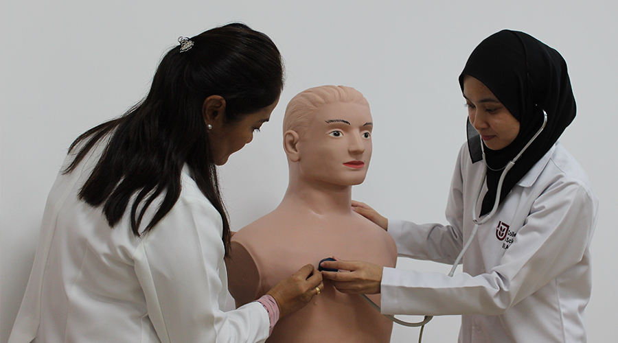 Nursing School in Asia | two female nursing students doll mannequin stethoscope asian Brunei JPMC College of Health Sciences classroom hearing heartbeat lab coat