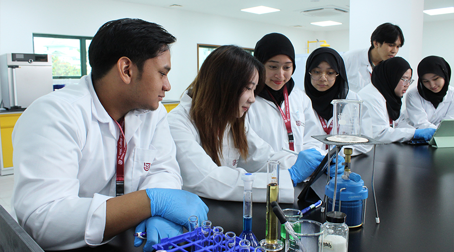 Nursing School Brunei | Group of male female JCHS students glasses lab coat name tags classroom bench Bunsen burner wire gauze beaker test tubes flasks studying tablets smiling