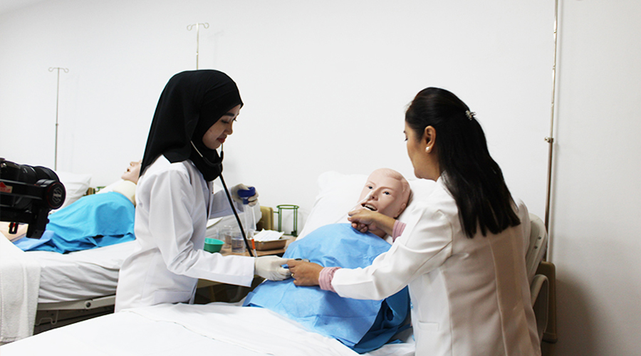 Nursing School Brunei | Female student lab coat stethoscope nursing manikins hospital bed side table learning