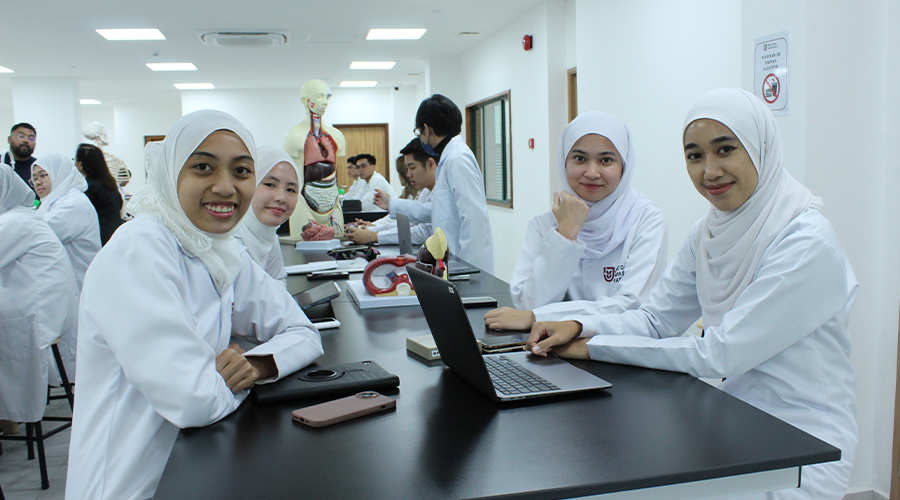 Nursing in Brunei | male and female JCHS students classroom bench anatomy model phones and laptops no food sign lights and windows door stool lab coat