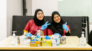 Nursing in Brunei | JCHS female students smiling school uniform name tag blue gloves clinical equipment table and chair thumbs up