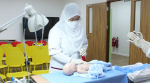 Nursing School Brunei | JCHS female nursing student classroom clinical practice baby table latex gloves table and chairs TV and door