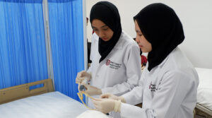 Nursing School Brunei | Two female nursing students with black hijab and latex gloves using clinical equipment in a classroom ward near hospital bed