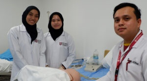 Nursing Course in Brunei | Male and female nursing students with labcoats, black hijabs and lanyard with red pen nursing mannikin on hospital bed clinical practice classroom ward